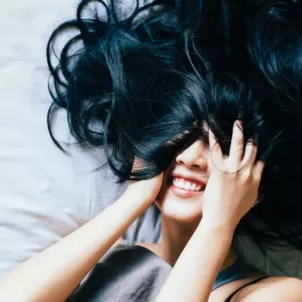 Woman with black hair smiling in bed with hair covering her eyes