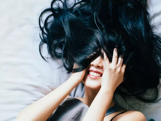 Woman with black hair smiling in bed with hair covering her eyes