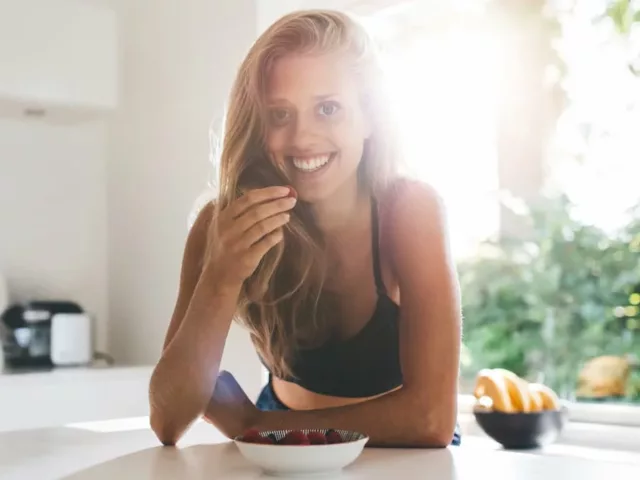 Fit blonde woman smiling in kitchen eating berries and healthy food to lose weight