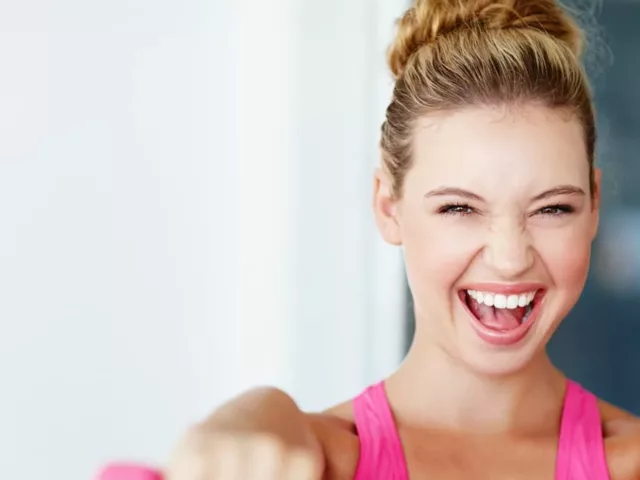 Woman in pink athletic gear smiling while raising dumbbells because she reached her happy weight