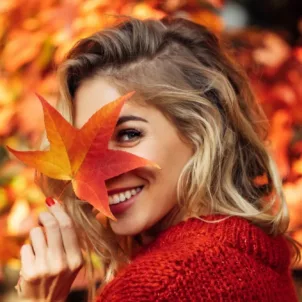 Woman in red sweater holding leaf in front of her face amidst the fall foliage