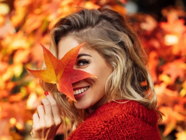 Woman in red sweater holding leaf in front of her face amidst the fall foliage