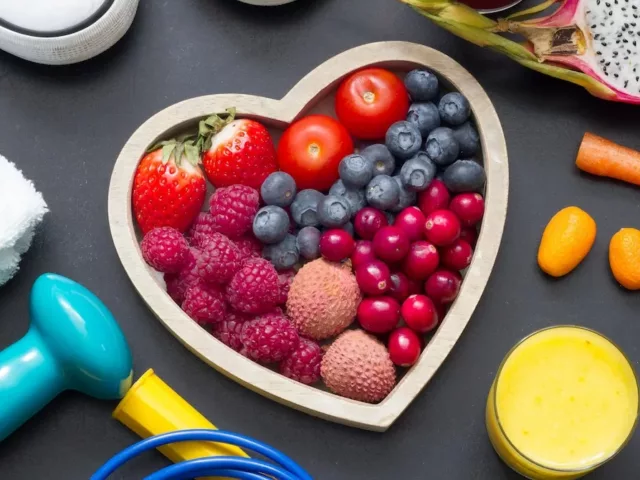 Heart-shaped bowl with berries, lychee, and tomatoes amidst fitness gear and orange juice on slate background