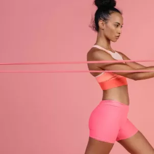 Beautiful woman in bright athletic gear pulling a band in front of pink background