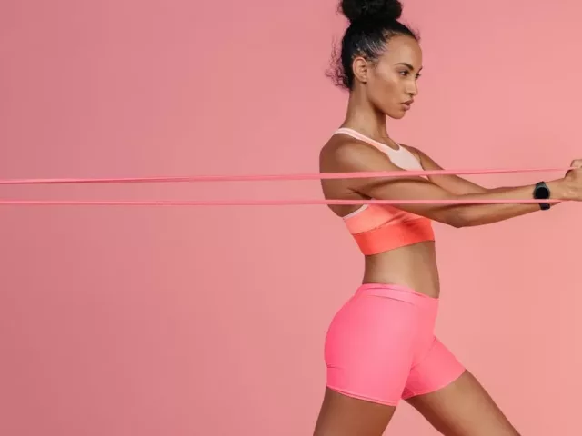 Beautiful woman in bright athletic gear pulling a band in front of pink background