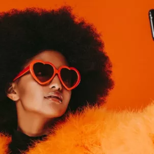 Woman wearing orange fur and heart-shaped sunglasses taking a selfie in front of red background