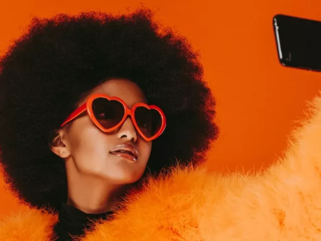 Woman wearing orange fur and heart-shaped sunglasses taking a selfie in front of red background