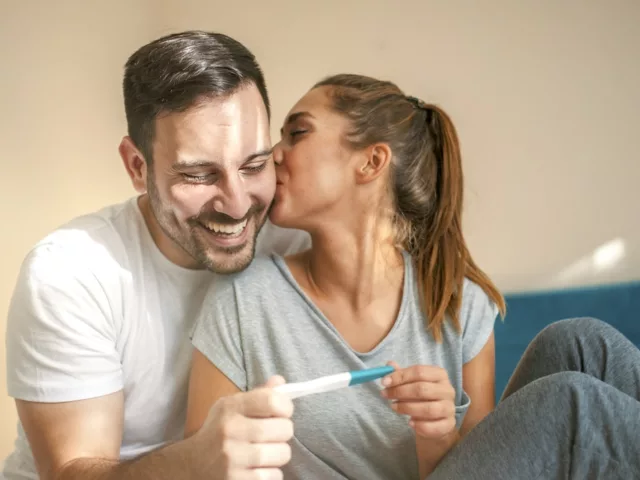 Happy couple in bed with positive pregnancy test; pregnancy tips concept