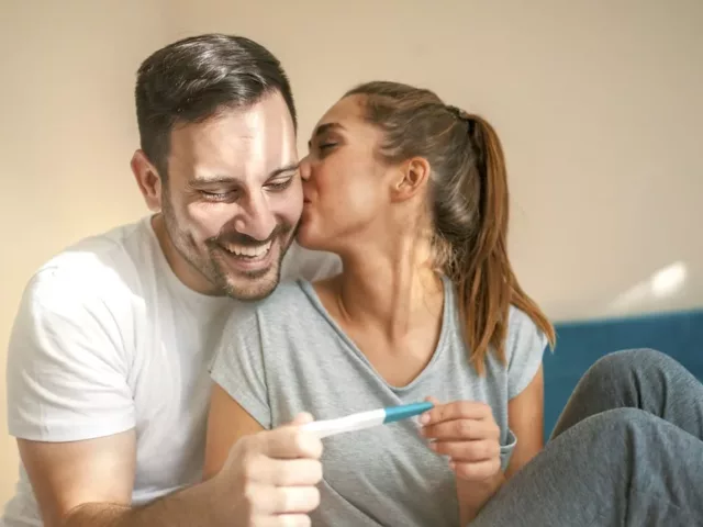 Happy couple in bed with positive pregnancy test; pregnancy tips concept