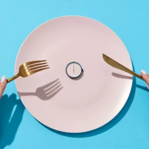 Woman's hands holding gold fork and knife in front of pink plate with a clock on it, signifying intermittent fasting