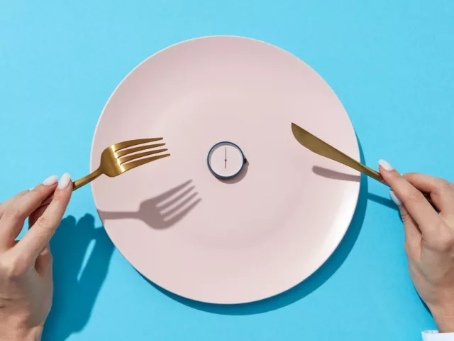 Woman's hands holding gold fork and knife in front of pink plate with a clock on it, signifying intermittent fasting
