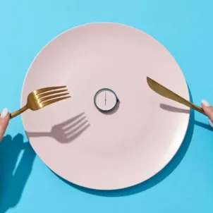 Woman's hands holding gold fork and knife in front of pink plate with a clock on it, signifying intermittent fasting