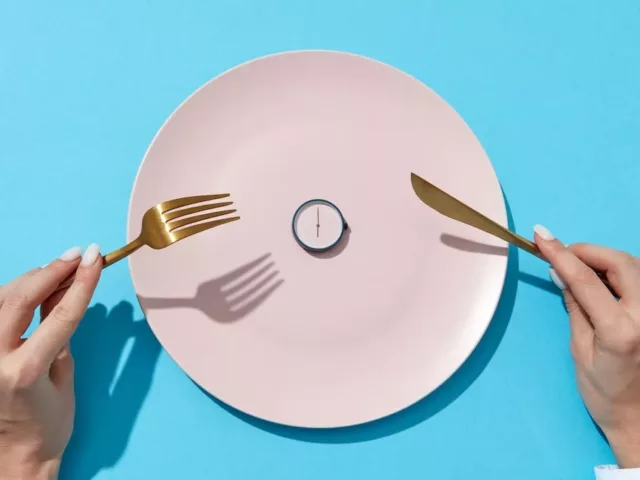 Woman's hands holding gold fork and knife in front of pink plate with a clock on it, signifying intermittent fasting