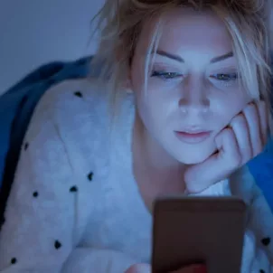 Woman faced with blue light emitted from her phone screen while under the covers in bed