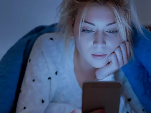 Woman faced with blue light emitted from her phone screen while under the covers in bed