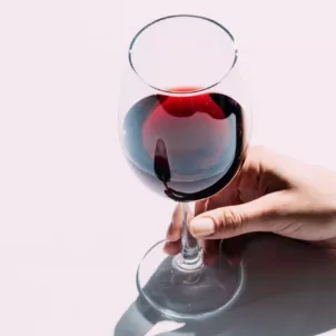 Woman holding a glass of red wine by the stem on top of pink background