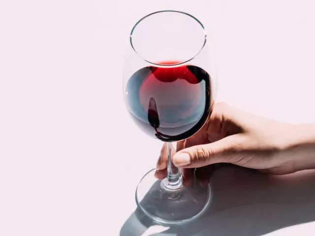 Woman holding a glass of red wine by the stem on top of pink background