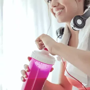 Woman with headphones and towel on shoulder drinking a protein powder shake from pink bottle