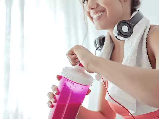 Woman with headphones and towel on shoulder drinking a protein powder shake from pink bottle