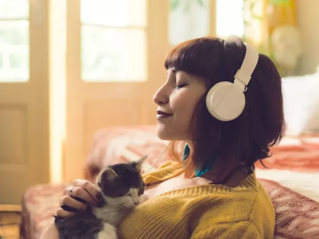 Woman on floor in bedroom holding kitten listening to meditation app on headphones