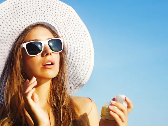 Woman in white sunhat and sunglasses applying sunscreen at the beach