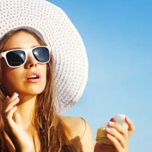 Woman in white sunhat and sunglasses applying sunscreen at the beach