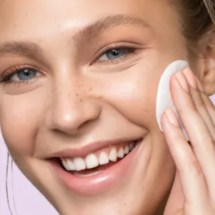 Young woman applying facial toner with a cotton pad, purple background