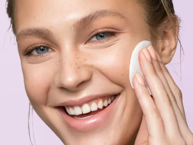 Young woman applying facial toner with a cotton pad, purple background