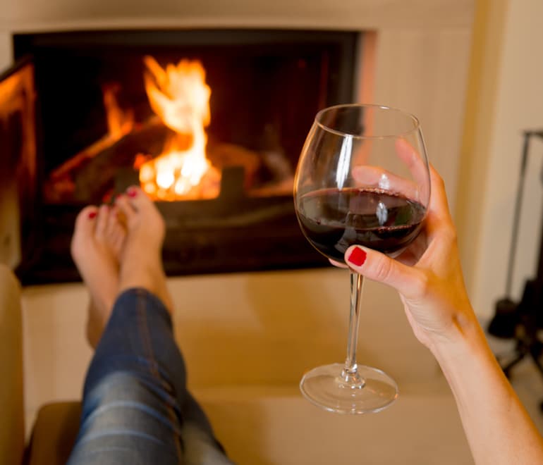 Woman holding a glass of red wine with legs kicked up at the fireplace for an anxiety-free Valentine's Day