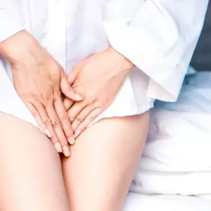 Woman in white shirt in front of bed with hands by her intimate areas to conceal vaginal odor