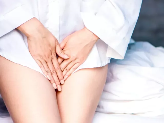 Woman in white shirt in front of bed with hands by her intimate areas to conceal vaginal odor