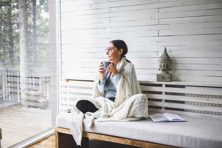 Woman sitting in her cozy reading nook drinking tea and looking out the window