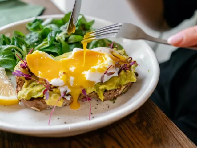 Woman eating a healthy breakfast for weight loss, cutting into an egg sandwich with avocado and leafy greens