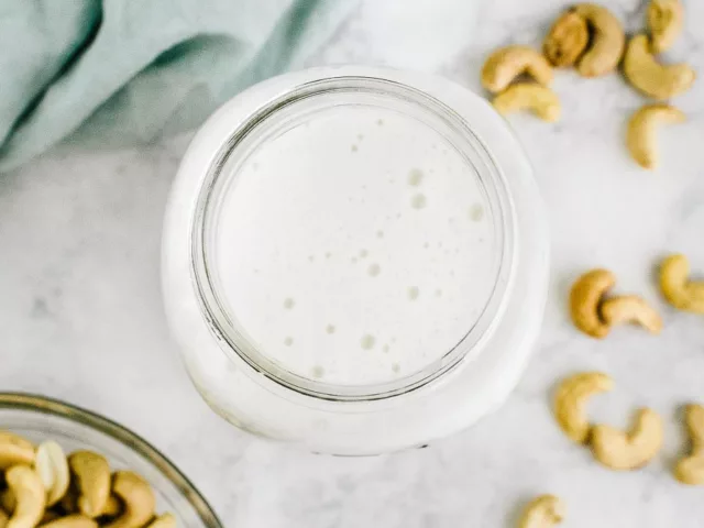 Homemade nut milk in a glass jar next to bowl of cashews and blue kitchen cloth