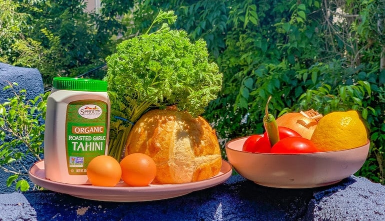 Ingredients for a traditional shakshuka recipe including onion, tomato, parsley, eggs, bread, and tahini paste