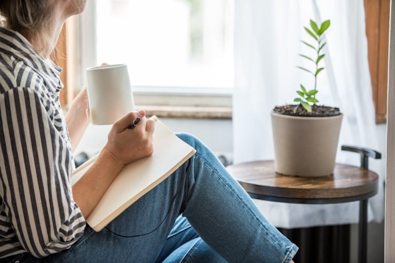 Woman writing in a journal with a mug of coffee by her window, discovering how to achieve work-life balance