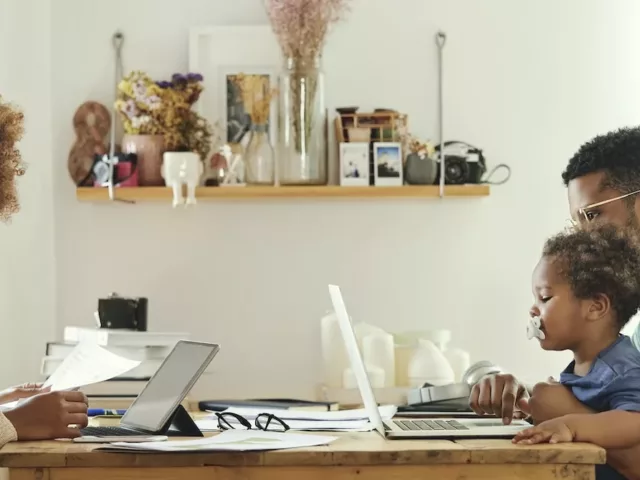 Young parents working from home on laptops in kitchen while the father holds a baby, needing to find work-life balance