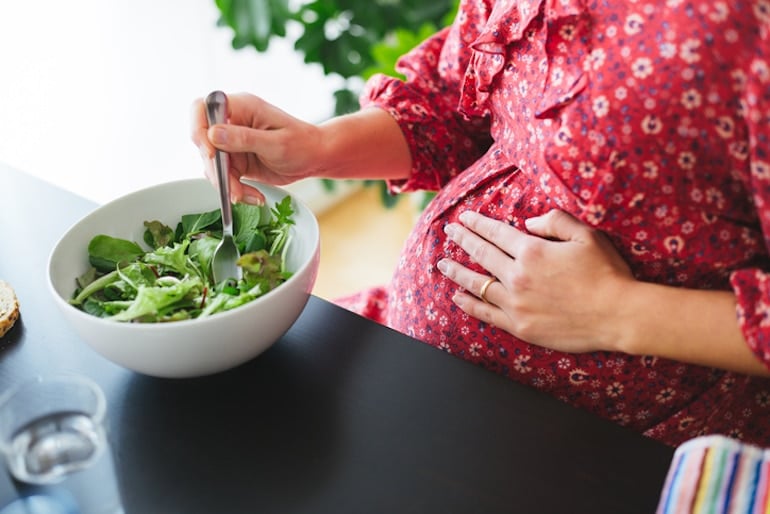 Pregnant woman eating a salad at the dinner table touching her belly, hoping to get the right nutrients through supplements and diet