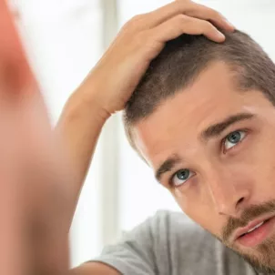 Young man touching hair in mirror concerned about men's hair loss and what causes it