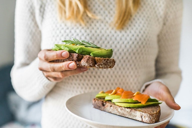 Blonde woman holding toast with salmon and avocado, two of the best foods for cramp relief and reducing PMS pain