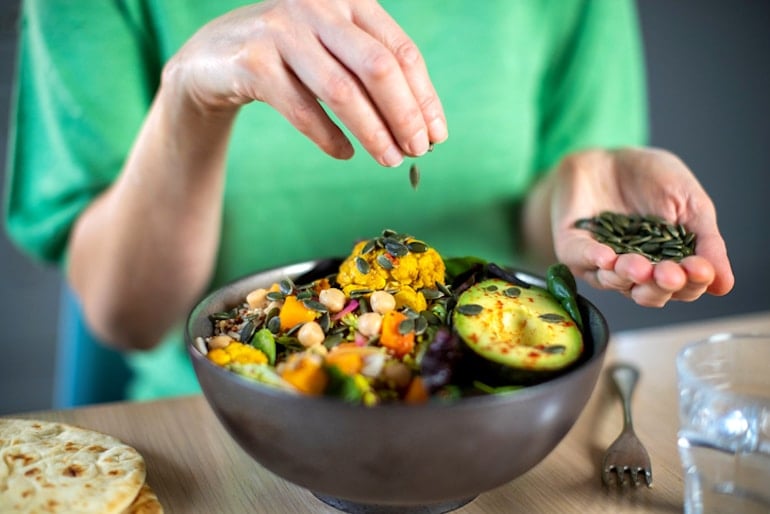 Woman dropping pumpkin seeds into her whole foods lunch bowl, illustrating healthy bio-individual diet concept