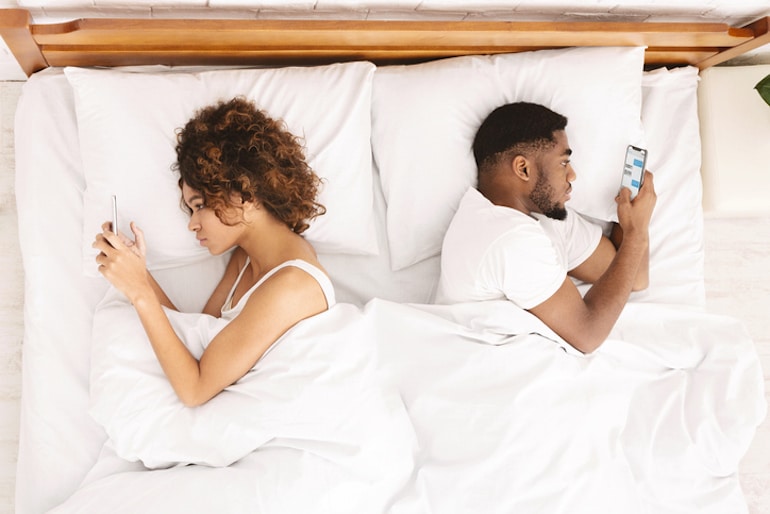 Young couple in bed with backs turned toward each other because they're busy on their phones, showing how your social life suffers from internet and phone use