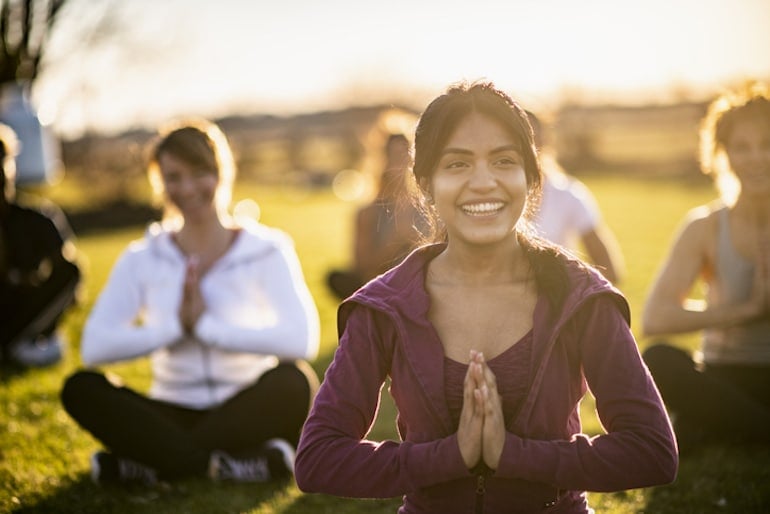 Group of people meditating in park, with girl smiling from the brain benefits of meditation