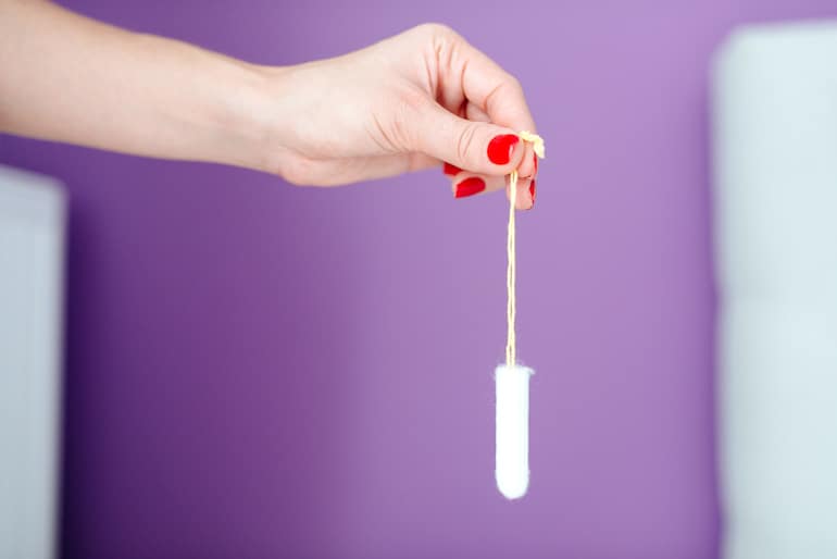 Woman holding an organic tampon by the string in front of a purple wall