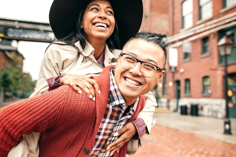 Woman wearing a large hat on friend's piggyback outdoors, smiling because hats don't make you lose hair or make your hair thinner