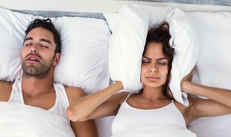 Woman blocking her ears with a pillow because her boyfriend is snoring and sleeping with his mouth open