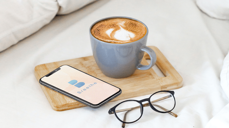 Phone with the Breethe meditation app, cup of coffee, and glasses on a bed