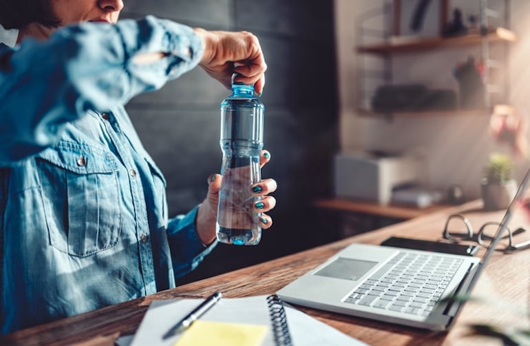 Woman opening bottle of water at her desk, since hydrating can improve your focus at work and prevent side effects of dehydration; a reason why it's important to stay hydrated