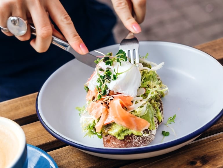 Woman cutting into salmon avocado toast with a poached egg, a healthy high-fat meal that packs many health and beauty benefits