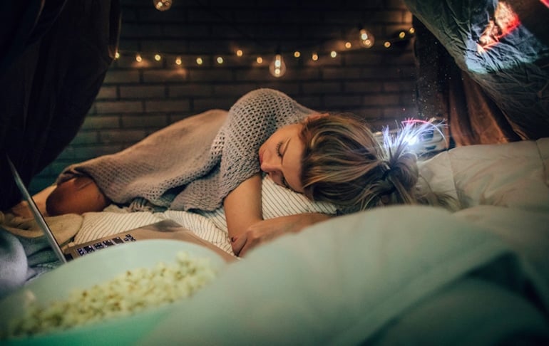 Woman watching movie in bed with midnight snacks, which can contribute to weight gain and poor sleep